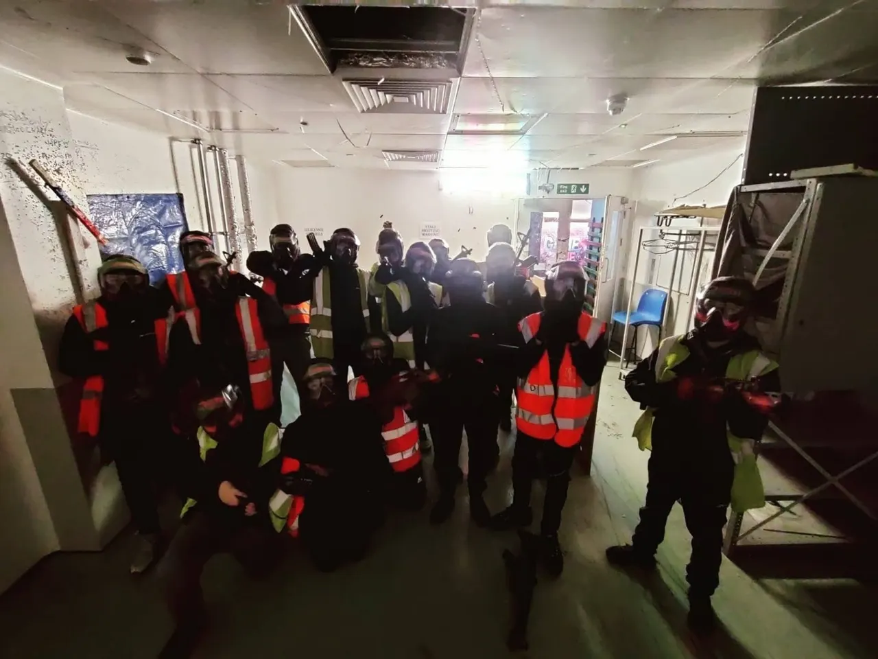 Large group of people in dark clothing and high-visibility vests gathered indoors during an airsoft activity session.