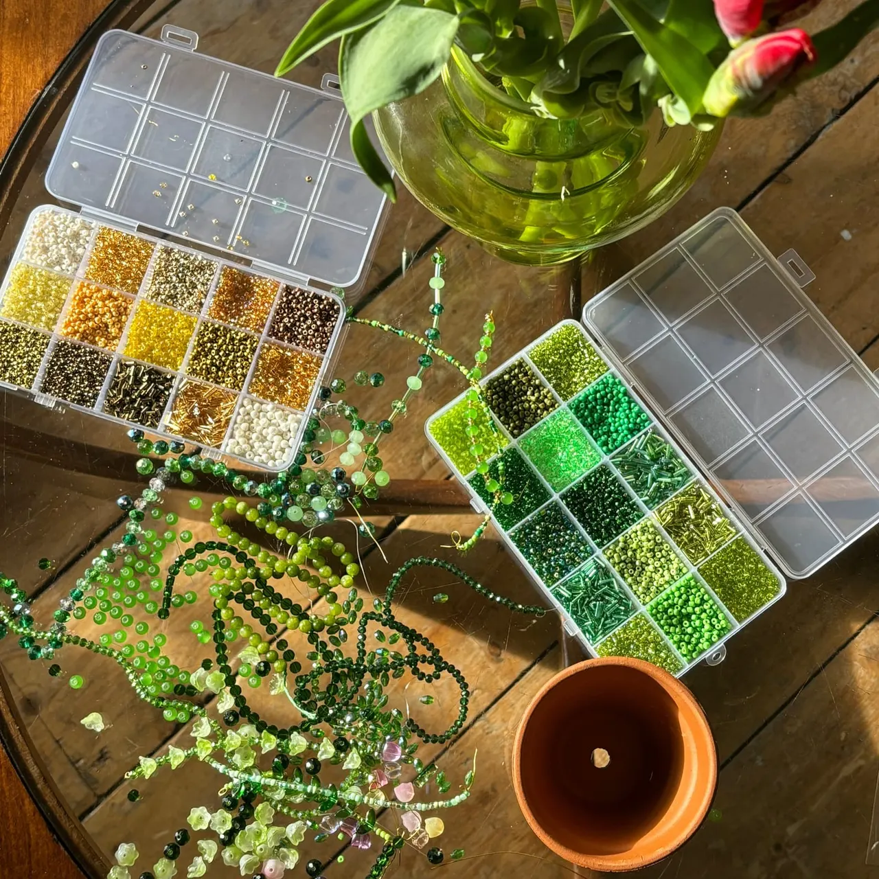 Overhead view of a beadmaking craft workspace with sorted beads in compartmentalised boxes, strung beaded jewellery, fresh flowers in water, and supplies on a wooden table.