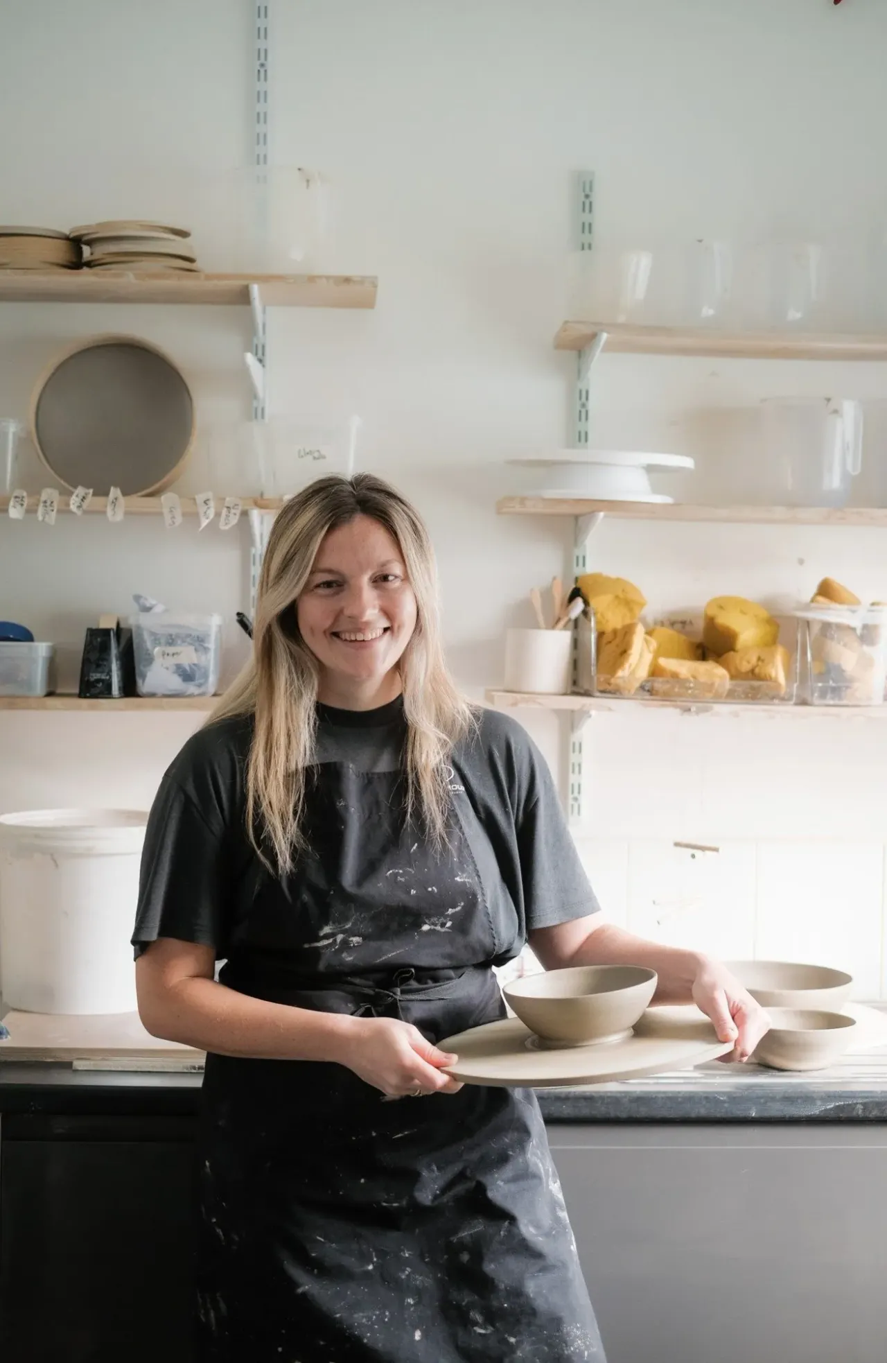 A woman in a black apron displays handmade ceramic bowls in a pottery studio workshop.