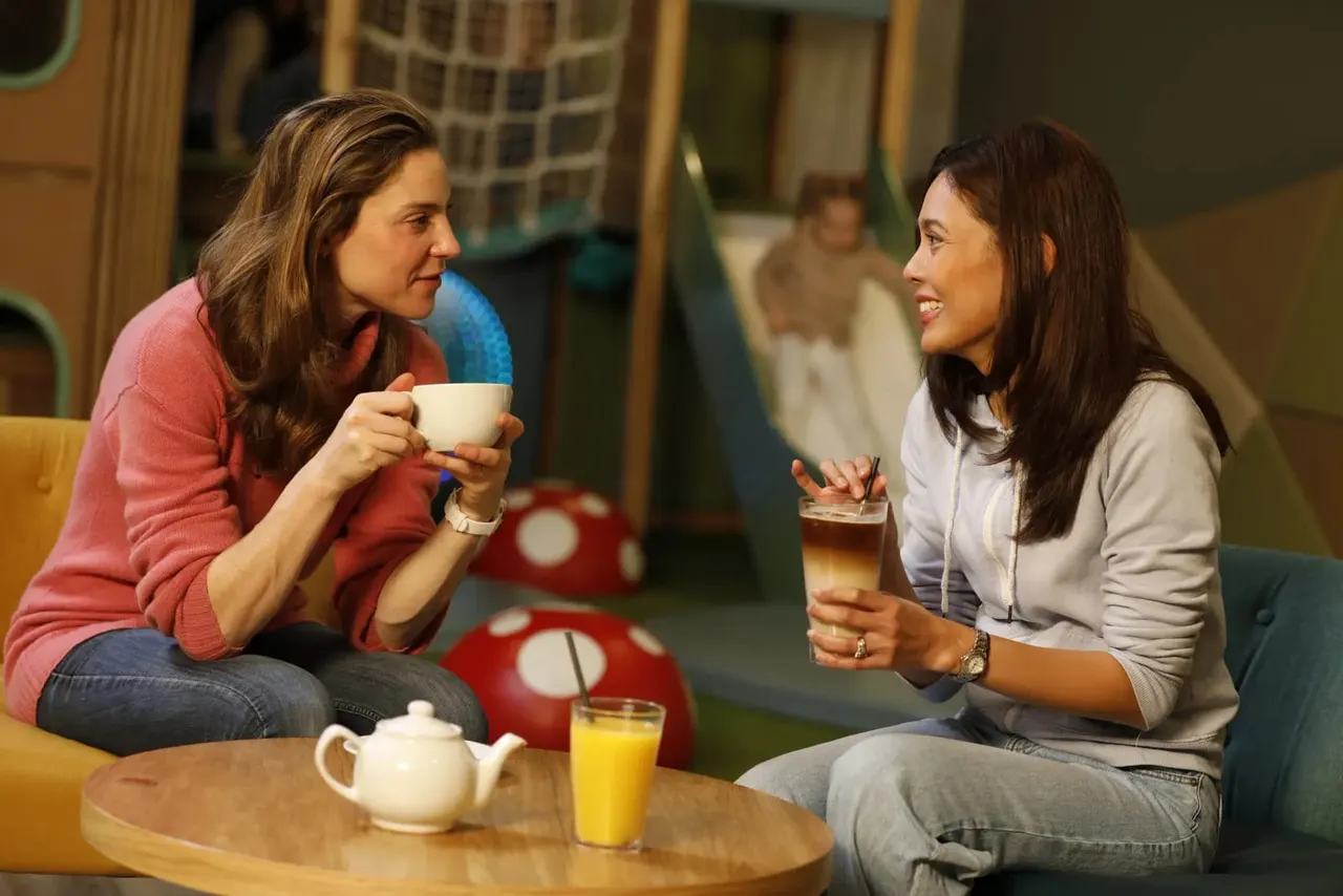 Two women enjoying drinks and conversation in a colourful indoor soft play area with seating and play equipment visible in the background.