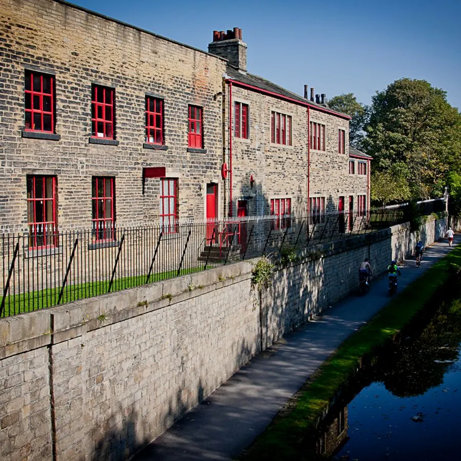 Stone mill building with red-framed windows beside a canal path where visitors walk in sunshine.