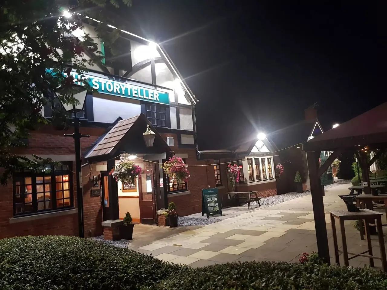 Nighttime view of a brick building with 'Storyteller' signage, hanging baskets of flowers, outdoor seating and paved courtyard area.