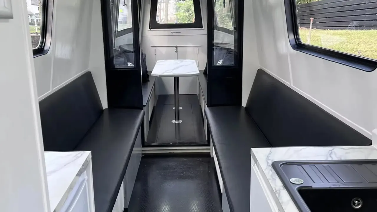 Interior of a passenger boat cabin with bench seating on either side, a central table, and access doors leading outside.
