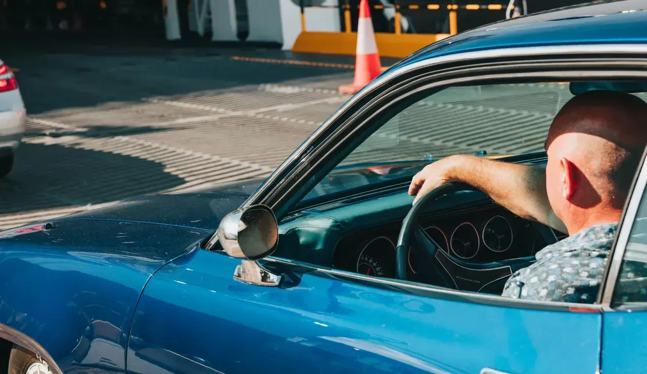 Man driving blue vintage convertible car at seaside leisure venue with traffic cones visible.