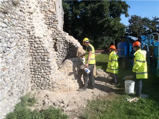 Workers in high-visibility clothing examining a historic stone wall ruins on a sunny day.