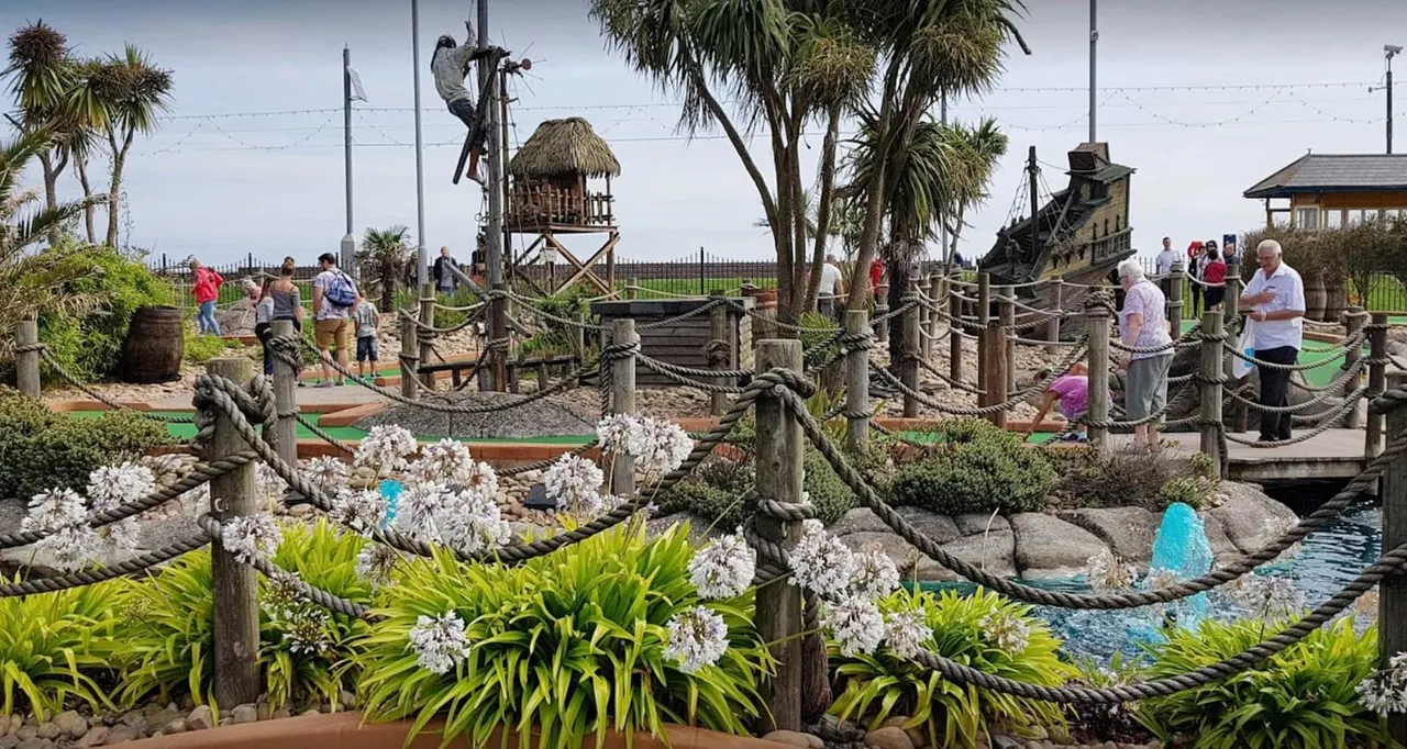 Families and visitors playing miniature golf on a pirate-themed course with rope railings, tropical plants, and ship-themed structures.