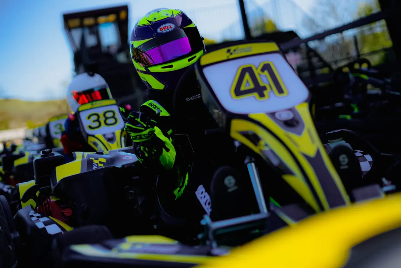 A kart driver in a neon yellow and black helmet sits in a bright yellow go-kart numbered 41, with other numbered karts lined up behind.
