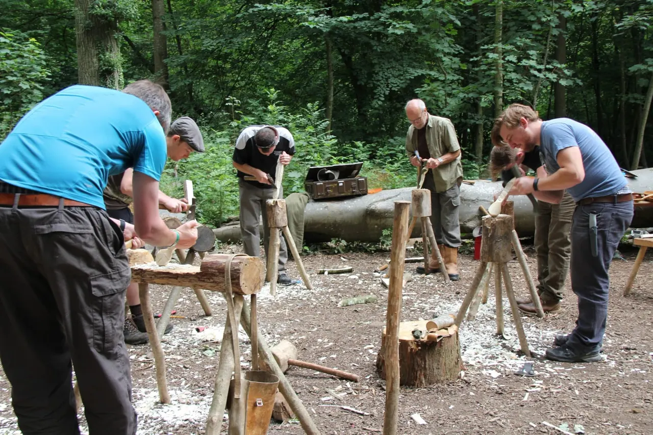 Group of adults engaged in woodworking activity, chiselling and shaping logs in a forest clearing.