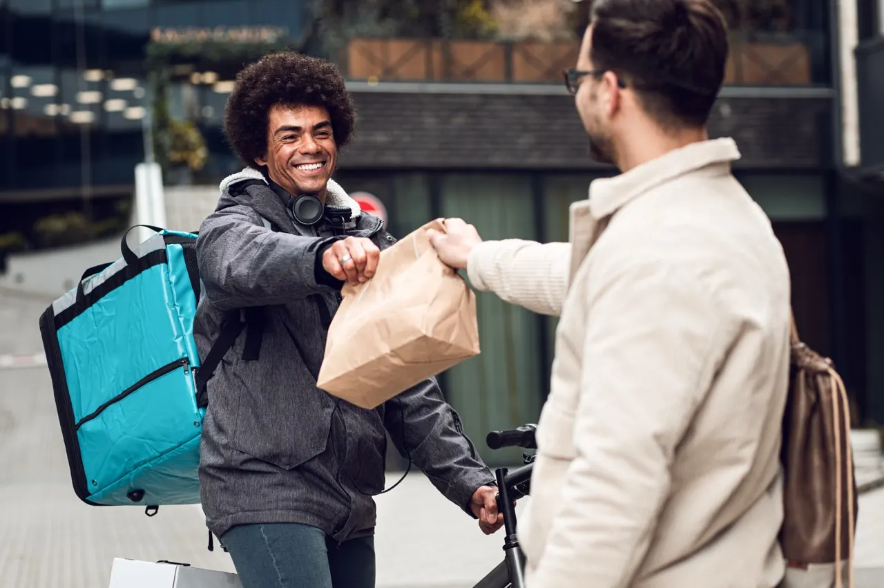 Delivery cyclist greeting customer with fist bump whilst holding food package on urban street.