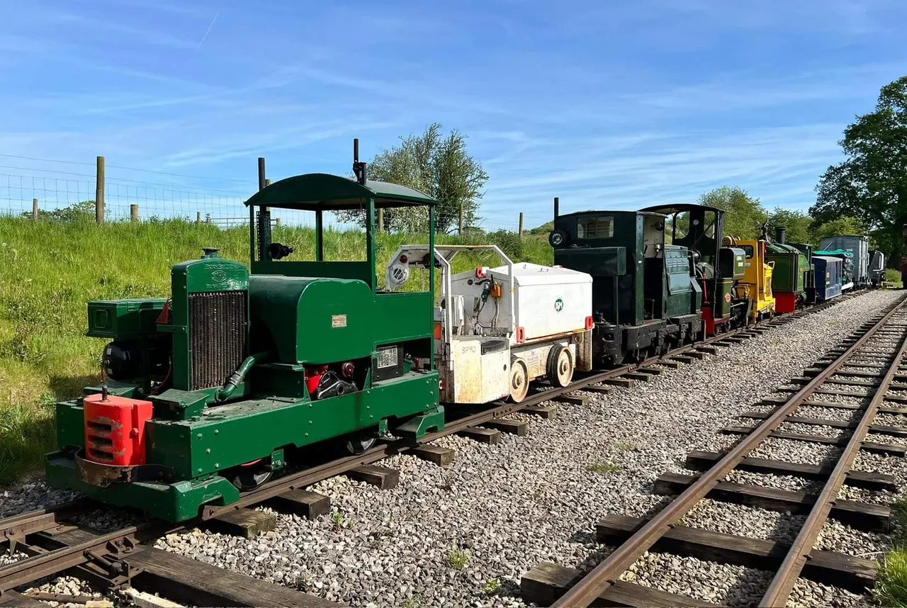 A green miniature steam locomotive pulling a line of heritage railway carriages along narrow-gauge tracks in a countryside setting.