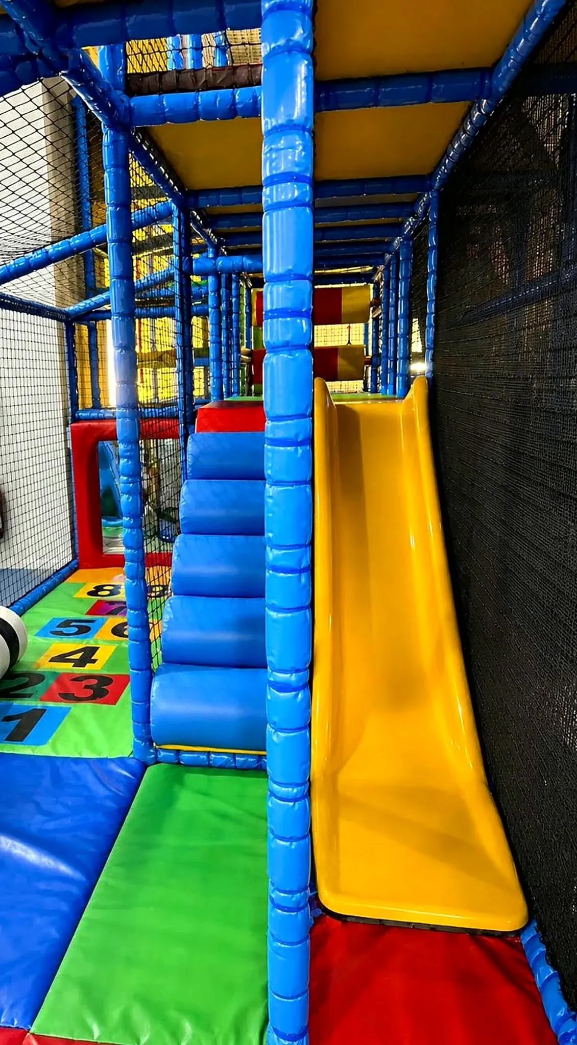 Interior view of a colourful indoor play structure with blue framework, yellow slide, and padded obstacles for children.