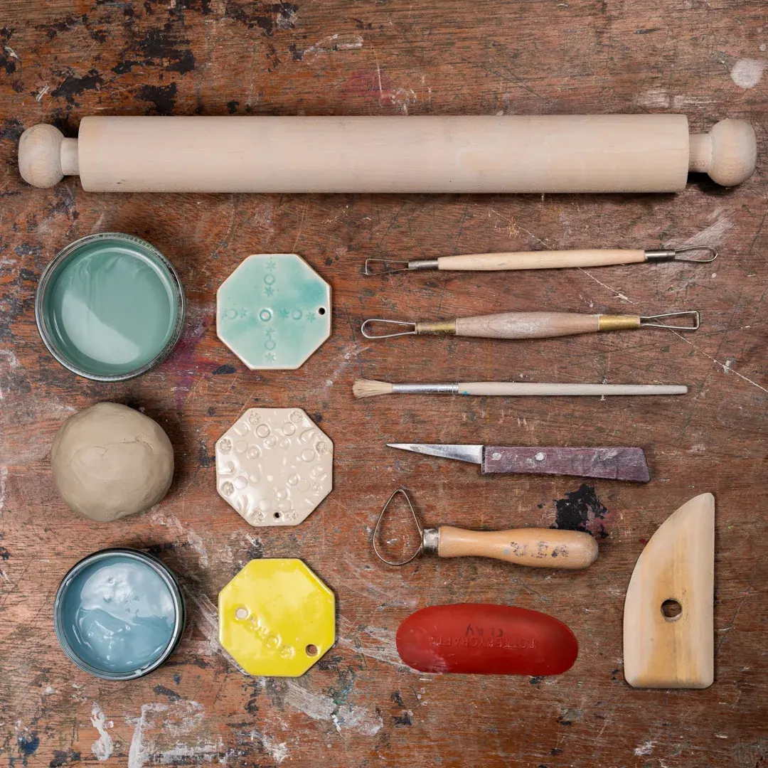 Pottery tools and materials arranged on a weathered wooden surface, including a rolling pin, clay, paint pots, sculpting tools and stamps.
