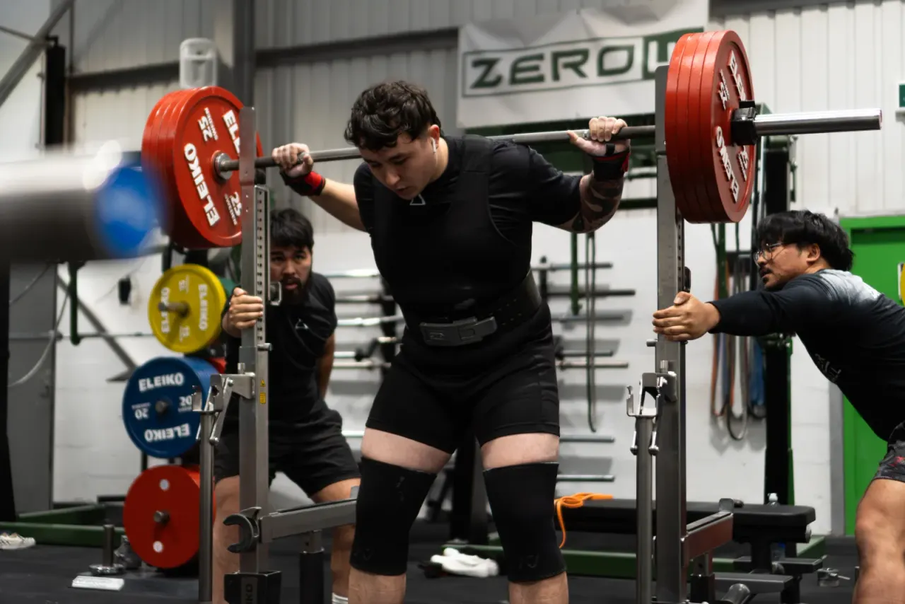 Weightlifter performing a barbell squat with red plates whilst spotted by two gym staff in an indoor gym facility.