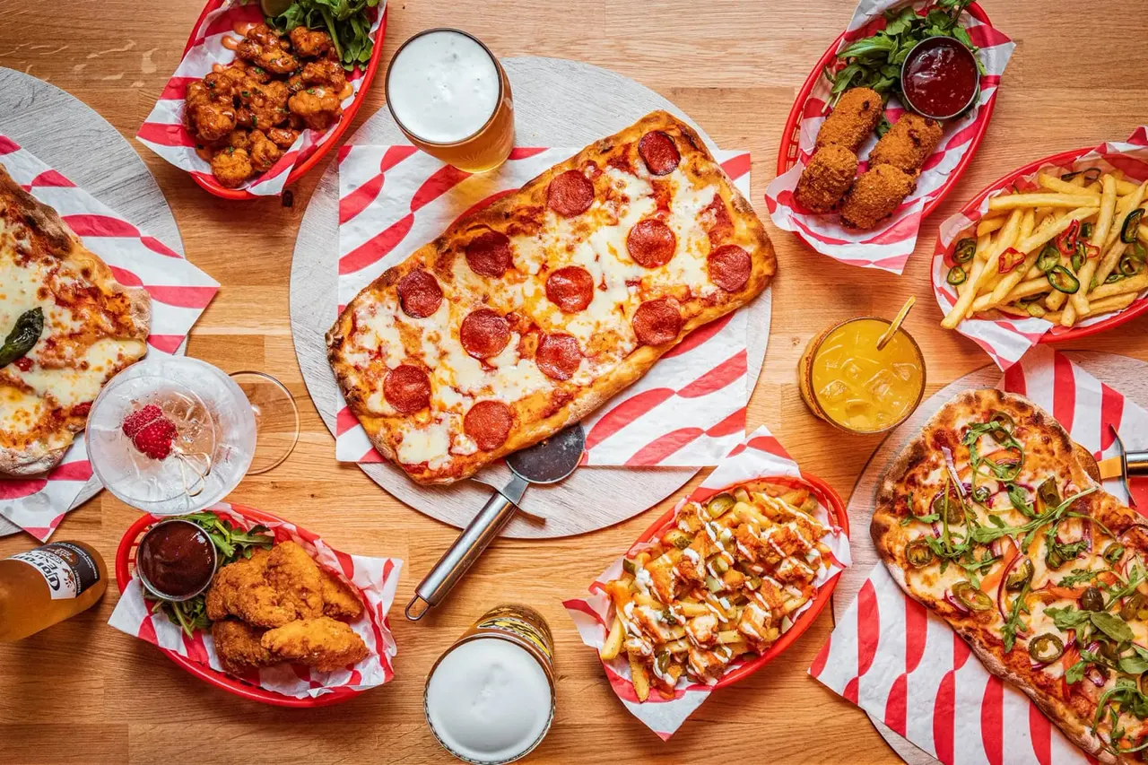 Overhead view of pizzas, fried chicken, chips and drinks arranged on a wooden table with red and white paper.