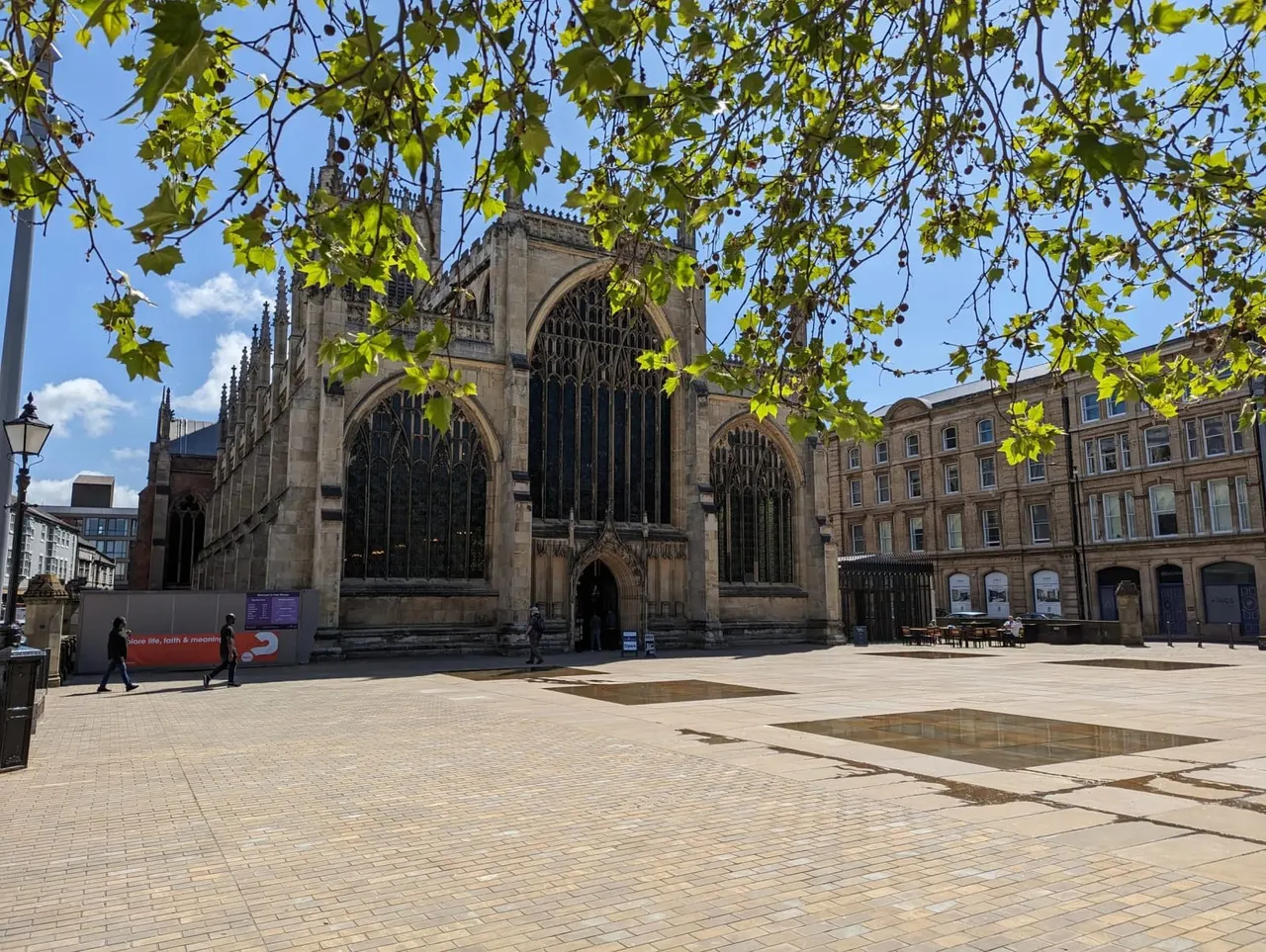 Gothic cathedral with large windows framed by hanging tree branches, people walking in sunny paved square below.