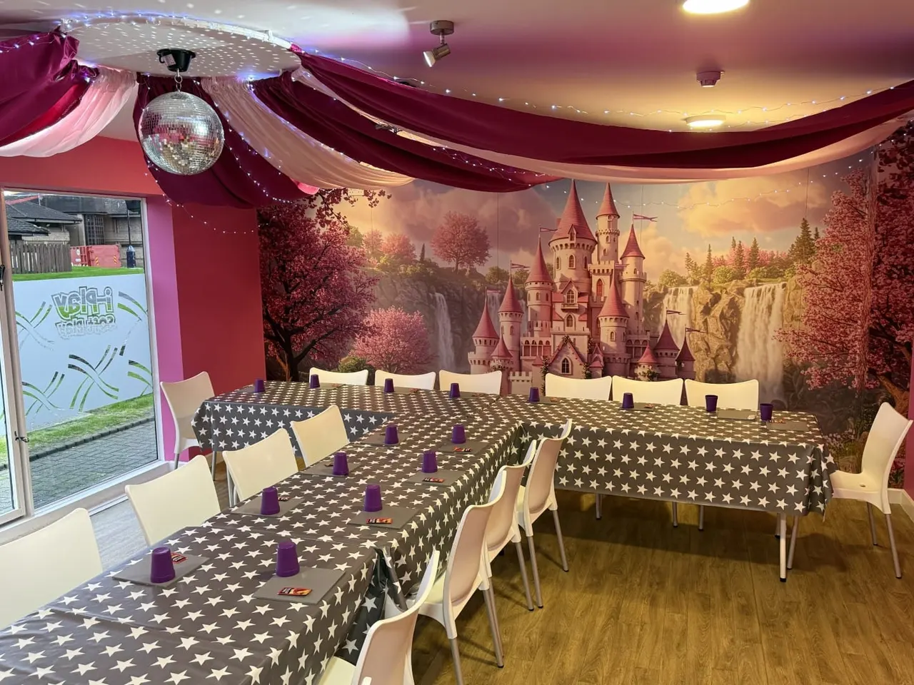 Decorated party room with long tables covered in star-patterned tablecloths, white chairs, purple candles, disco ball, draped fabric ceiling, and castle mural backdrop.
