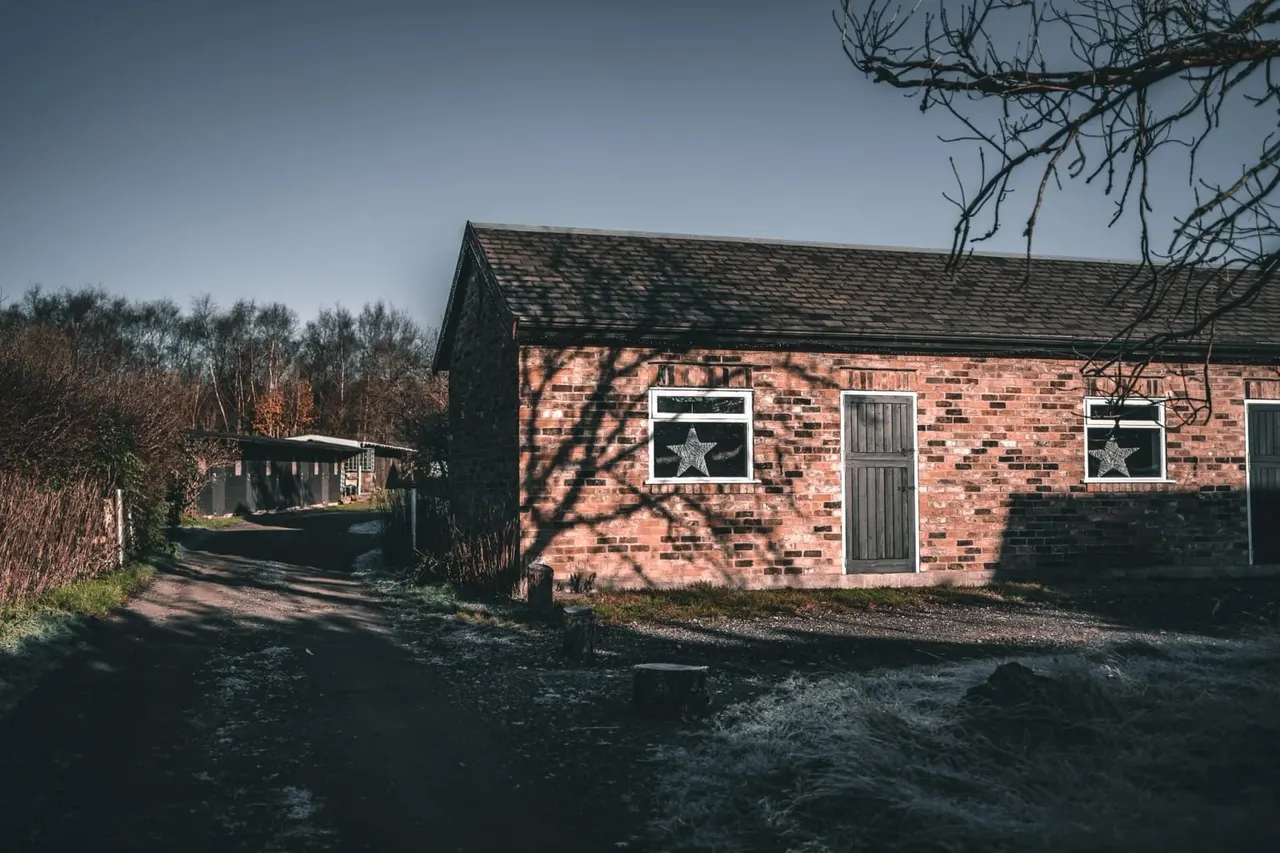 Red brick farm building with starlit window decorations, surrounded by bare winter trees and gravel courtyard.