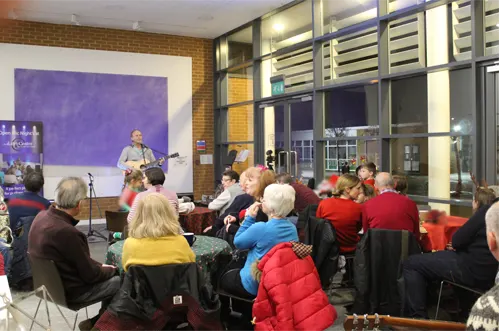 Audience members seated facing a musician performing with a guitar in front of a purple artwork.