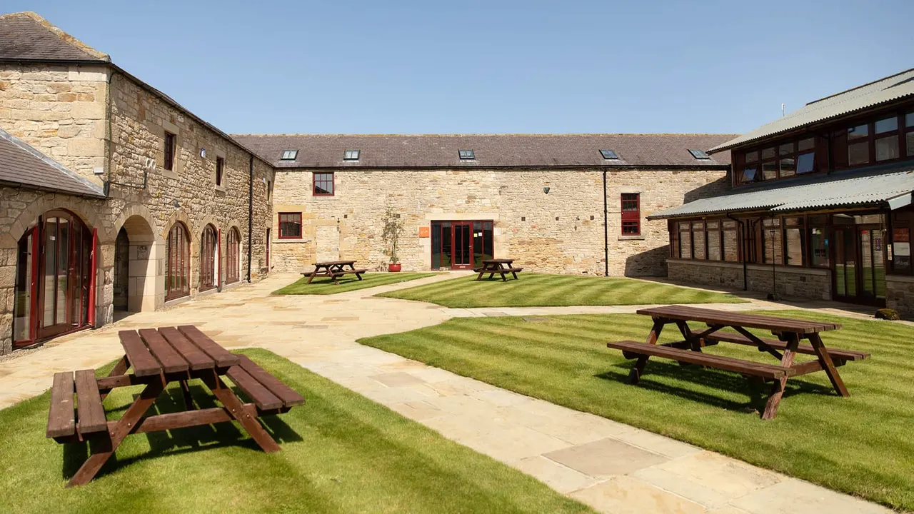 Stone courtyard with picnic tables on a lawn, surrounded by historic stone buildings with red doors.