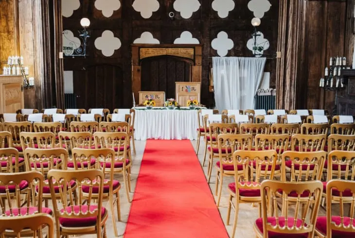 Wedding ceremony setup with red aisle, wooden chairs with burgundy cushions, and decorated stage with white backdrop.