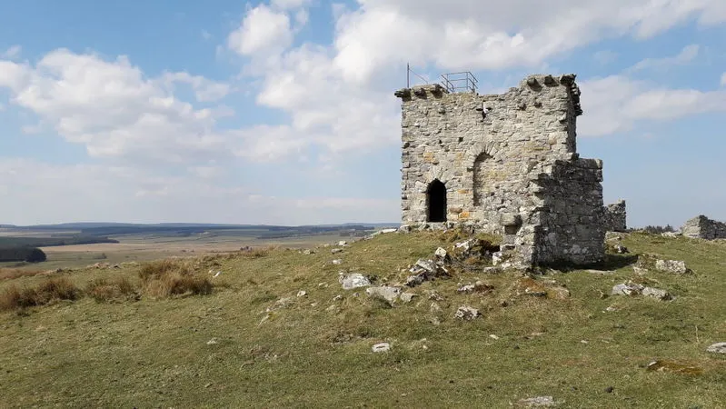 Ruined stone tower standing on moorland with expansive valley landscape and cloudy sky in background.