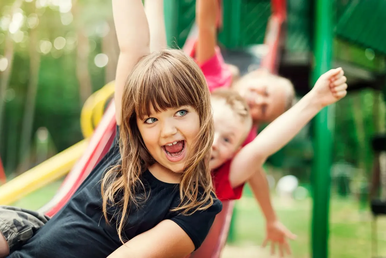 Young child on a red slide, laughing with arm raised, other children visible behind on playground equipment.