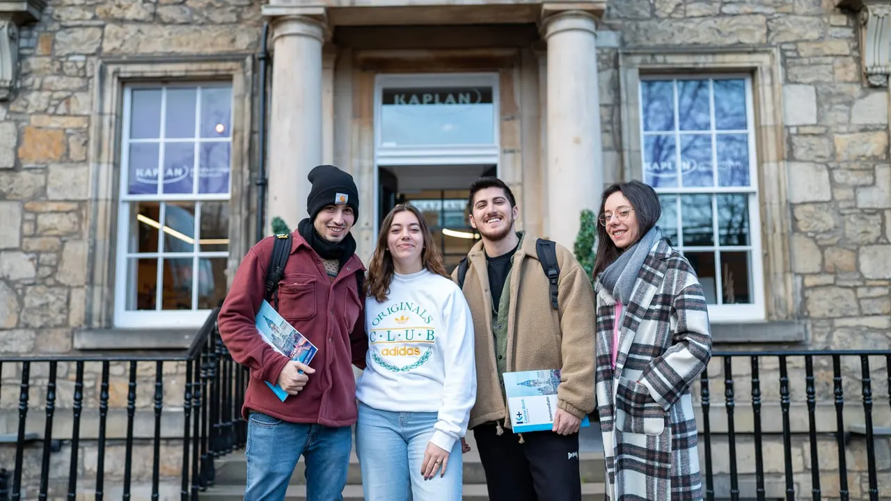 Four young adults standing together outside a stone building entrance marked "Kaplan", holding leaflets and smiling at the camera.