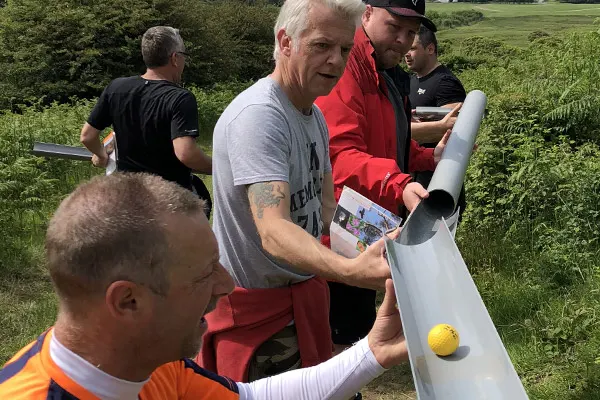 A group of adults examining a white pipe or tube outdoors in a rural setting with green fields behind them.