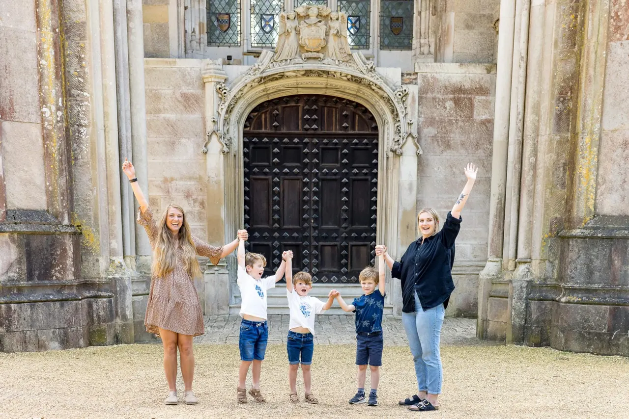 Two women and three young children hold hands in a line, celebrating in front of an ornate black studded door at a historic stone castle.