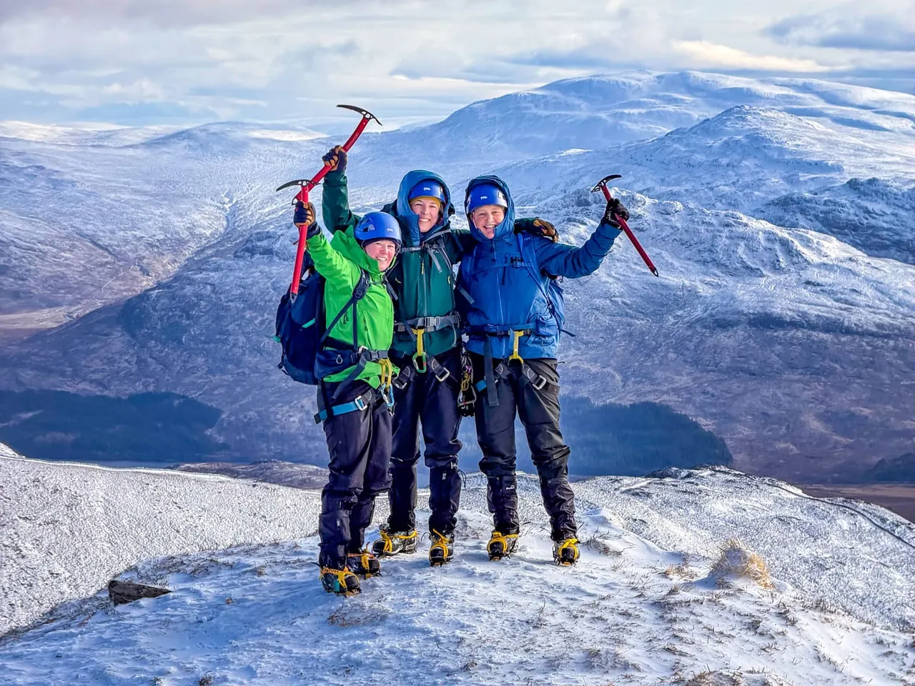 Four people in winter climbing gear and helmets celebrate on a snowy mountain summit, holding ice axes aloft with vast snow-covered peaks in the background.