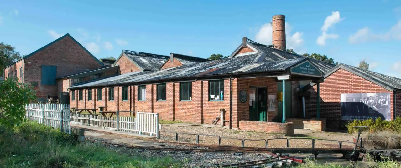 Historic red brick industrial buildings with chimneys set beside a narrow waterway under blue sky.