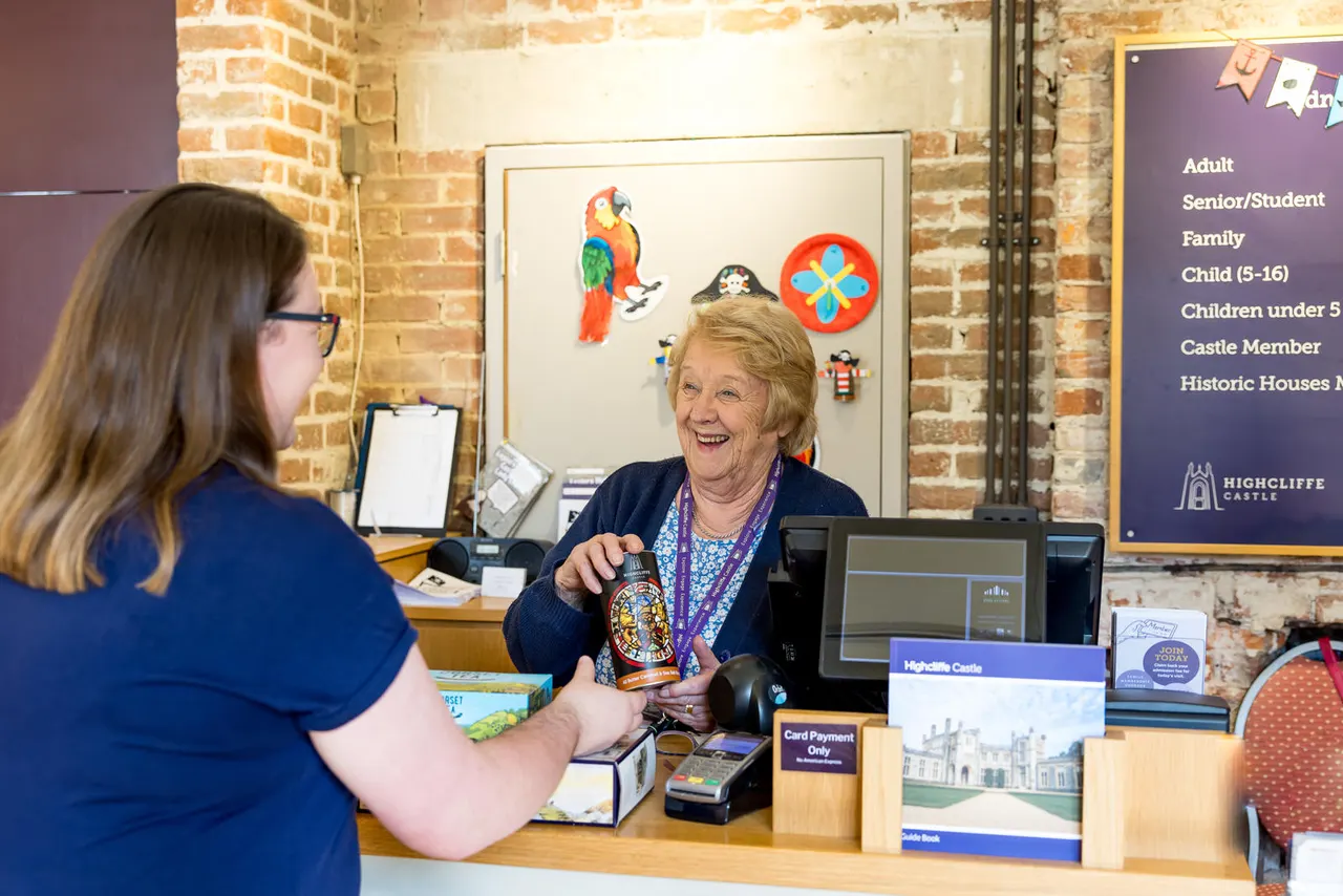 A staff member at a reception desk smiles whilst serving a visitor, holding up a souvenir bottle in front of a brick wall decorated with colourful artwork.