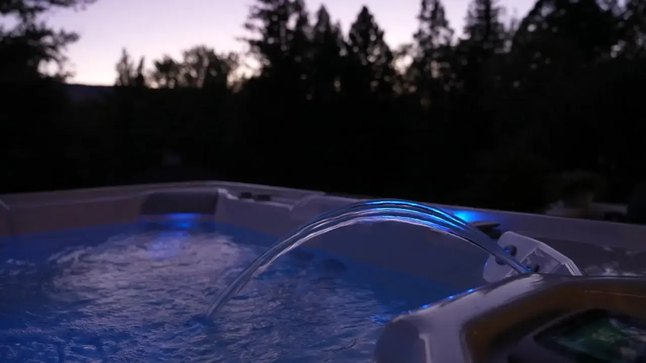 Hot tub with blue LED lighting at dusk, surrounded by dark conifer trees under twilight sky.