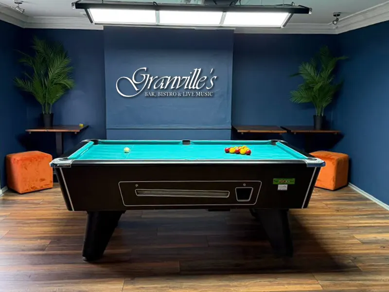 A pool table with turquoise felt and coloured balls in a blue-walled games room with potted plants and orange seating.