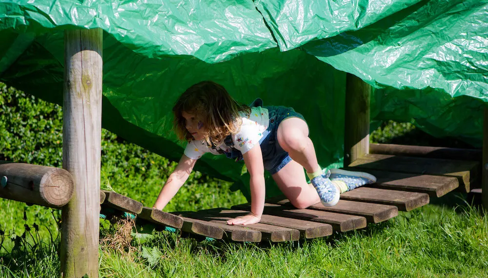 Child crawling through wooden tunnel structure covered with green netting at outdoor play area.