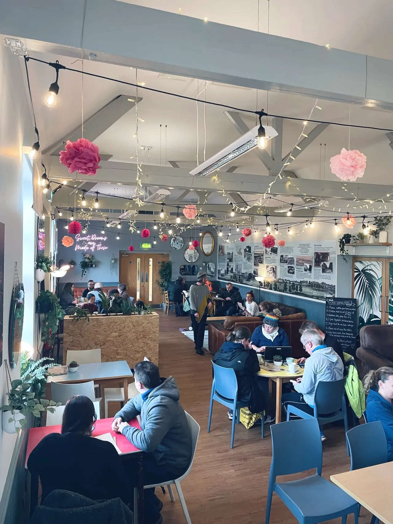 Interior of a busy café with visitors seated at tables and standing by a counter, decorated with hanging paper flowers and string lights.