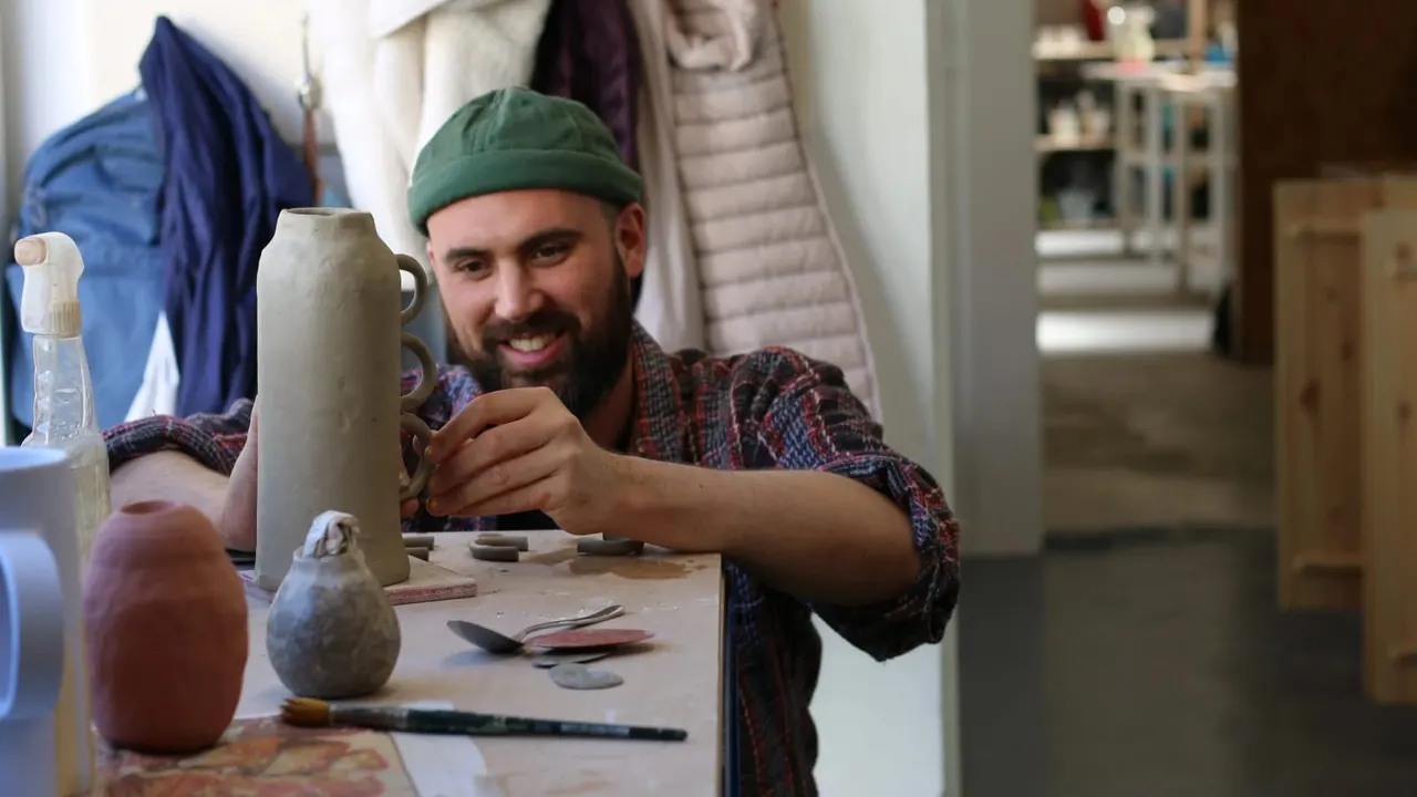 Man in green hat working on pottery at a table with clay vessels and sculpting tools.