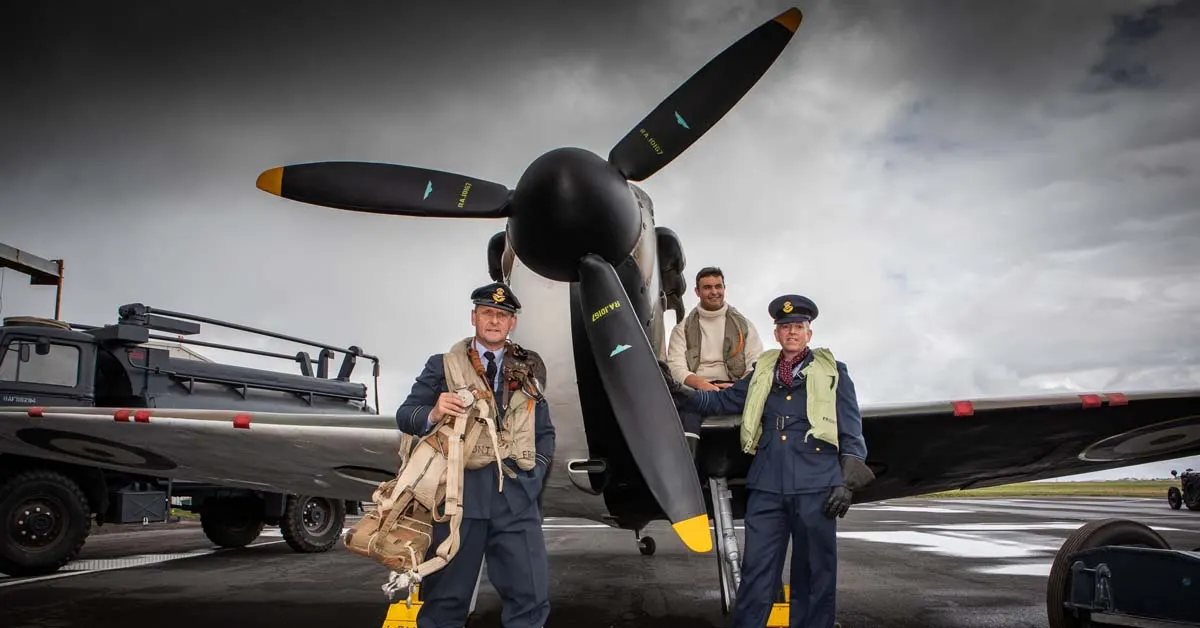 Three people in military uniform pose with a historic aircraft on an airfield under cloudy skies.