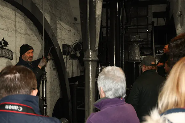 A guide leads a group of visitors on a tour inside an industrial building with brick walls and metal pipework.