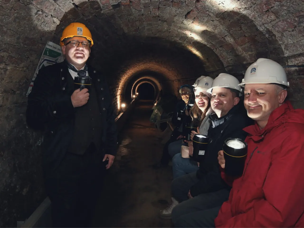 A guide in an orange hard hat leads a group of visitors wearing white hard hats through an underground brick tunnel with arched ceiling and rusted pipes.