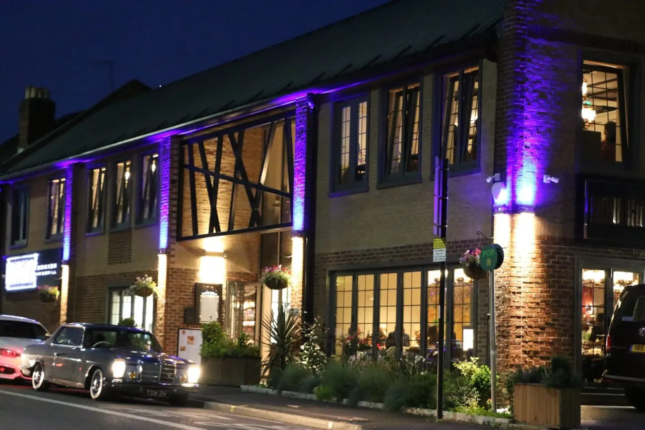 Brick building with purple and white accent lighting at night, featuring large windows and half-timbered sections, with parked car outside.
