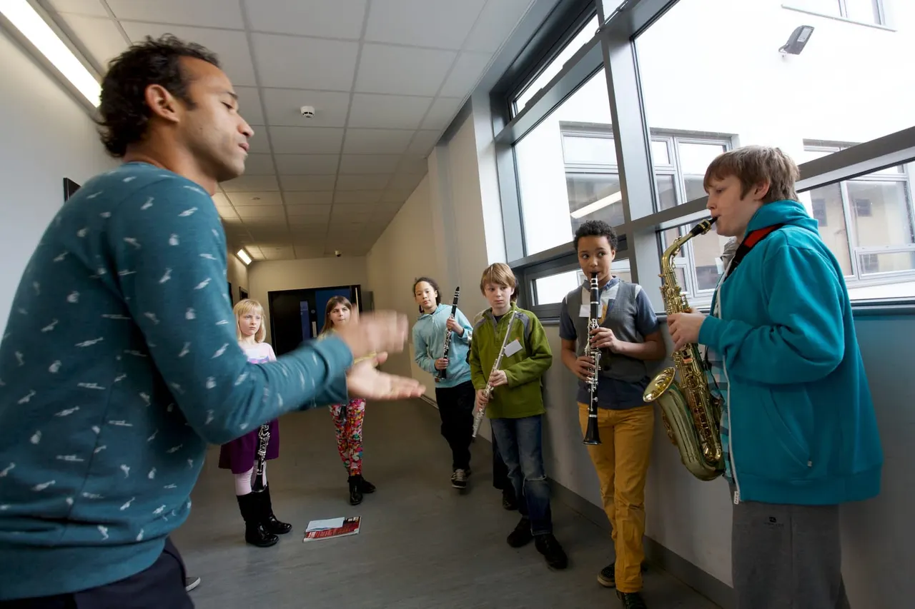 An adult music tutor conducts a group of children holding wind instruments in a bright indoor studio space.