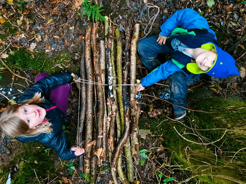 Two young children in bright jackets stand beside a moss-covered fallen tree trunk in a woodland setting.
