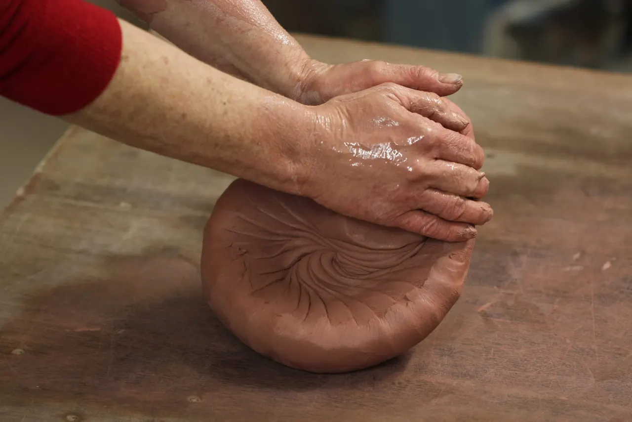 Adult's hands shaping wet clay on a wooden work surface during a pottery class.