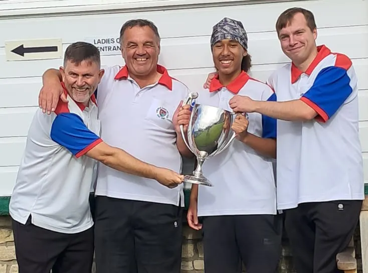 Four men in matching red and blue bowling shirts hold a trophy together, smiling at the camera.