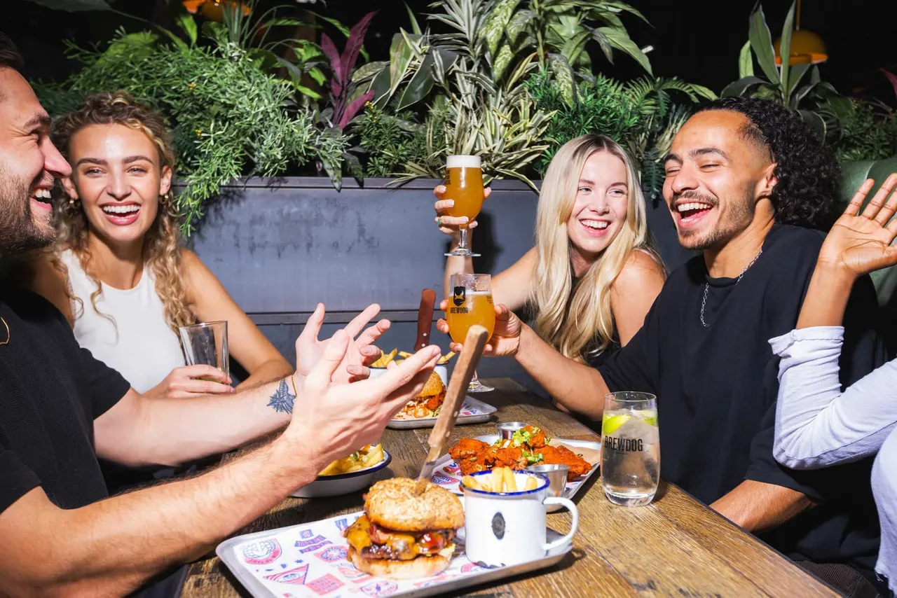 Group of five smiling people enjoying drinks and food at an outdoor night table surrounded by potted plants.