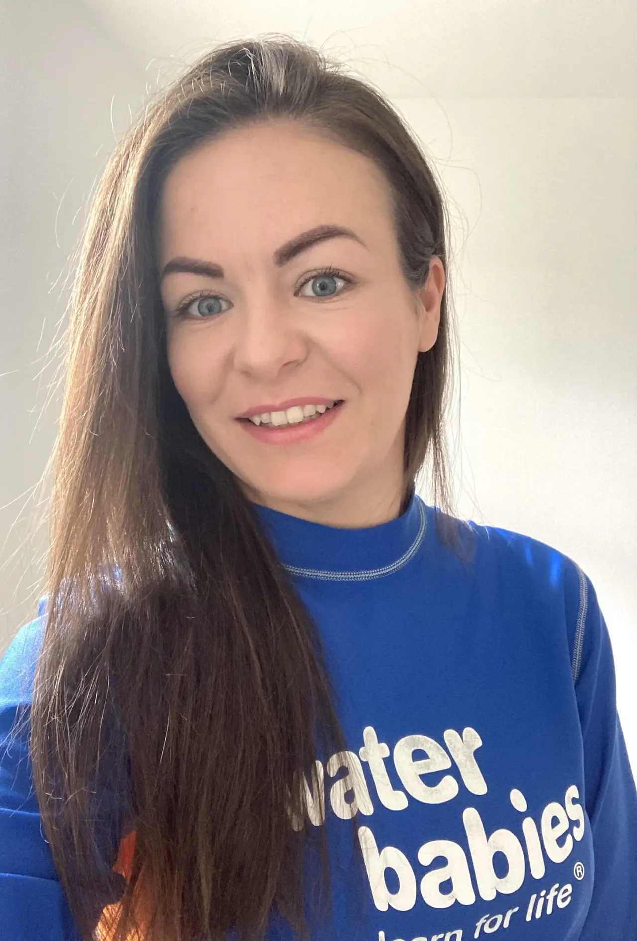 Woman wearing a blue Water Babies branded top, smiling at the camera against a white background.