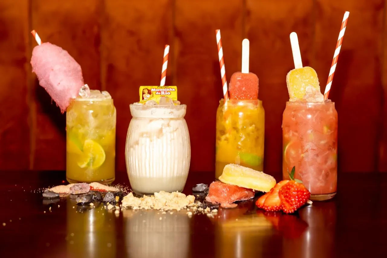 Four colourful cocktails with straws and garnishes arranged against an orange backdrop, with fresh fruit and sugar scattered on the surface.