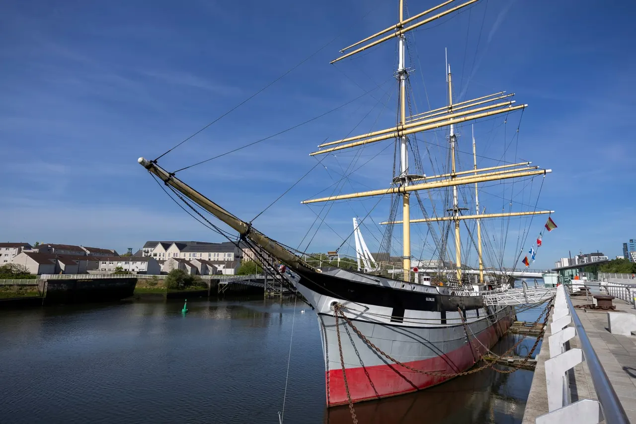 Historic tall ship moored at riverside docks with multiple masts and rigging visible against clear blue sky.