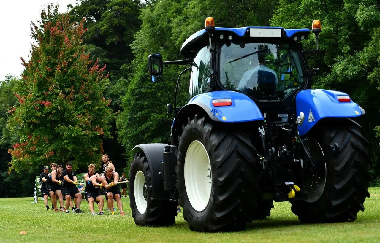 A large blue tractor with a bench trailer full of people on a grass field surrounded by trees.